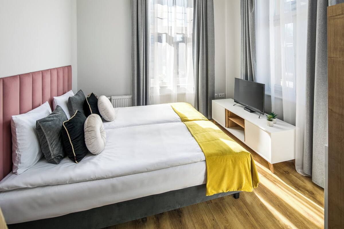 Una habitación de hotel moderna con una cama grande, un televisor sobre un mueble blanco y vistas a la ciudad a través de la ventana.