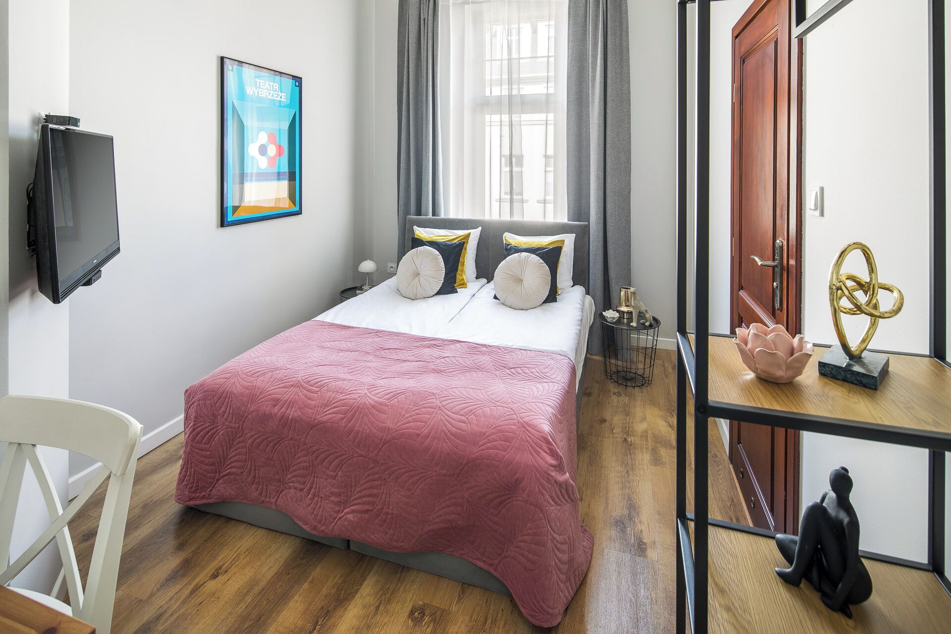 A bedroom with a bed, a TV, a window with curtains, a desk with a lamp, and a door.