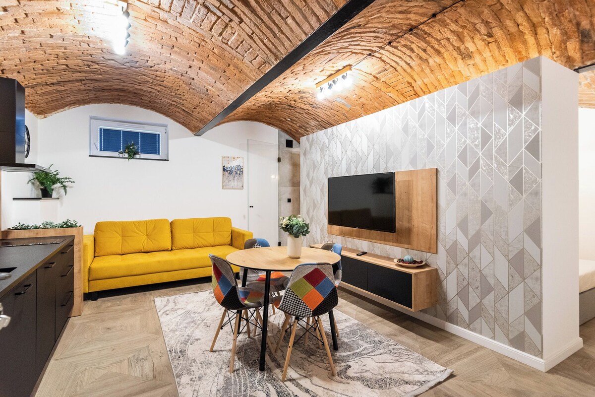 A modern living room with a yellow sofa, a round wooden table, and a TV mounted on the wall.