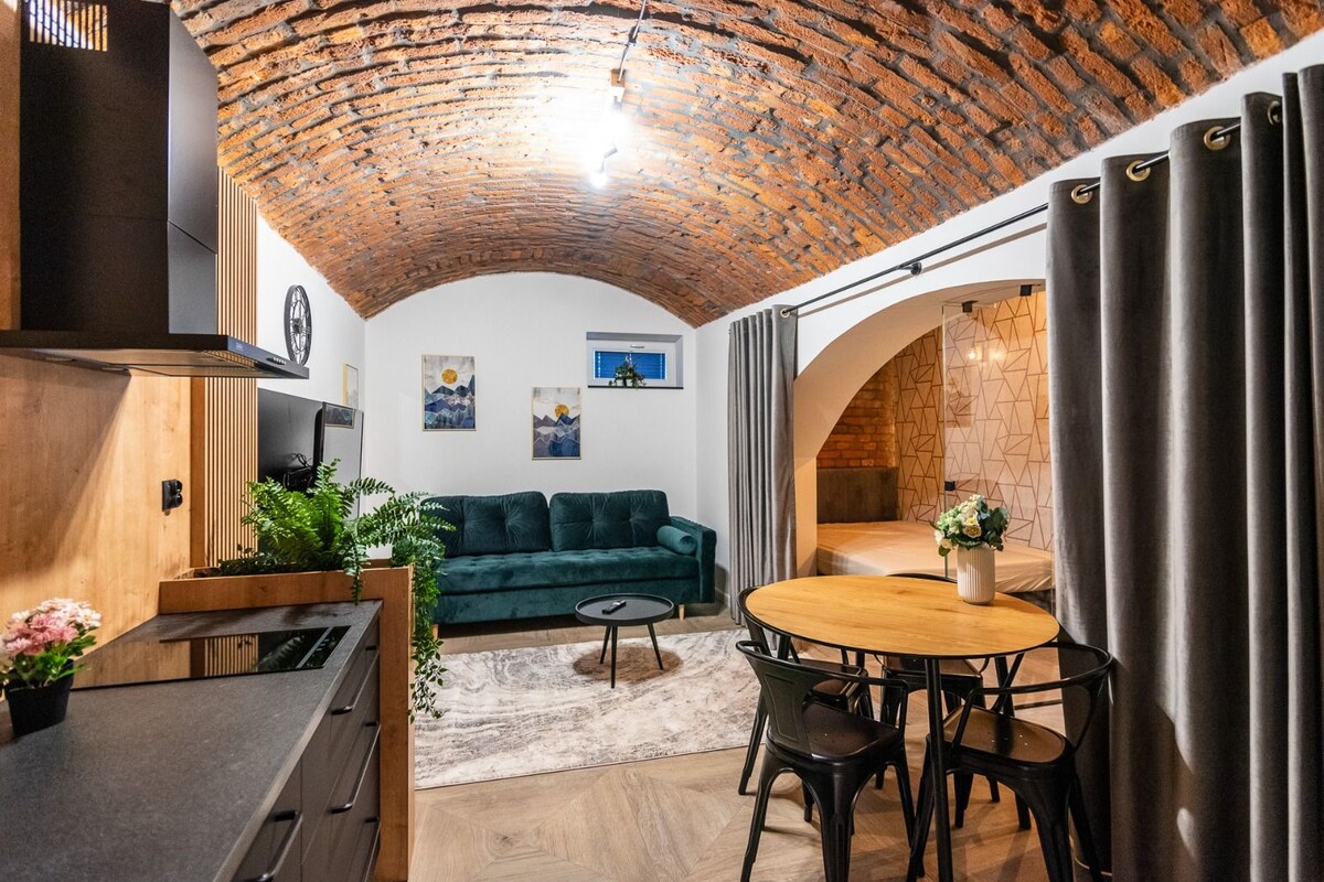 A cozy living area with a brick ceiling, a green sofa, a wooden dining table, and a kitchen area with a sink and cabinets.