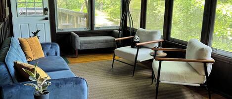 A cozy room with a blue sofa, a white armchair, and a wooden coffee table. There are large windows offering a view of greenery outside.