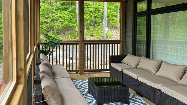 A covered outdoor seating area with two sofas and a coffee table, surrounded by wooden railings and large windows offering a view of a forest. - Twilight Pines - Four Bedroom Cottage, Sleeps 8 (Huntsville)