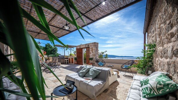 A covered outdoor seating area with stone flooring, a sofa, and a table with a laptop.