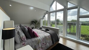Un dormitorio con una cama grande, una mesita de noche, una lámpara y vistas al exterior a través de amplias ventanas.