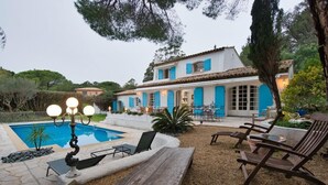 A pool area with lounge chairs and a table, surrounded by a garden and a building with blue shutters. - Villa with swimming pool walking distance from the beach (Ramatuelle)