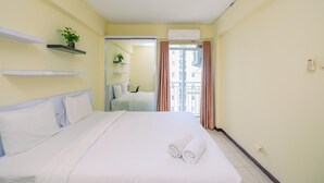 A hotel room with a bed, white bedding, a shelf with a plant, an air conditioning unit, and a view of a building through a window. - Cozy And Homey 3Br At Bogor Valley Apartment (Bogor)