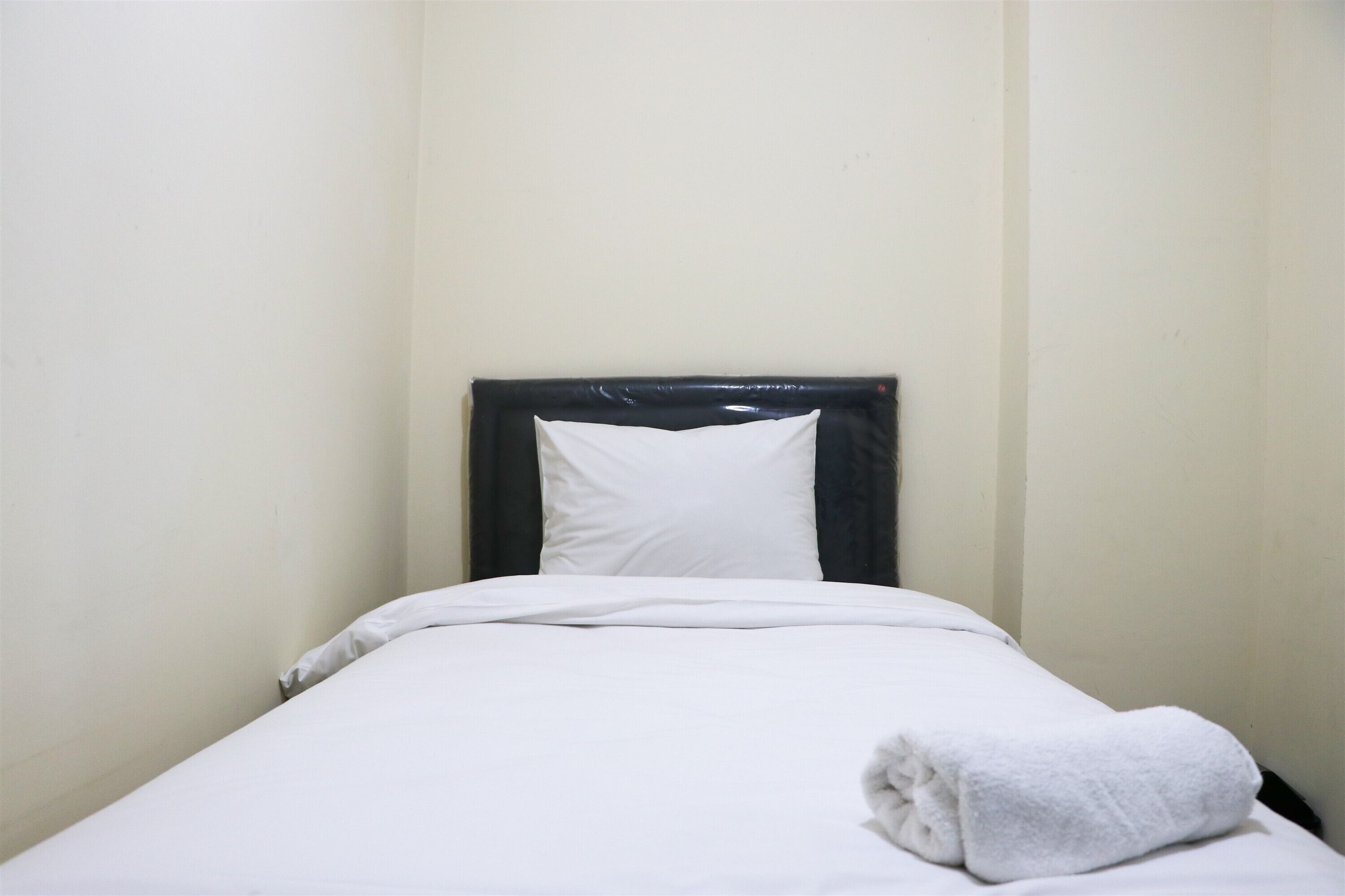 Ένα μονό κρεβάτι με ένα λευκό στρώμα και ένα λευκό μαξιλάρι, μια διπλωμένη λευκή πετσέτα στο πλάι και ένα μαύρο προσκέφαλο.