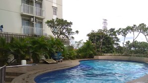 Una piscina de borde curvo, rodeada de follaje y un edificio con balcones.