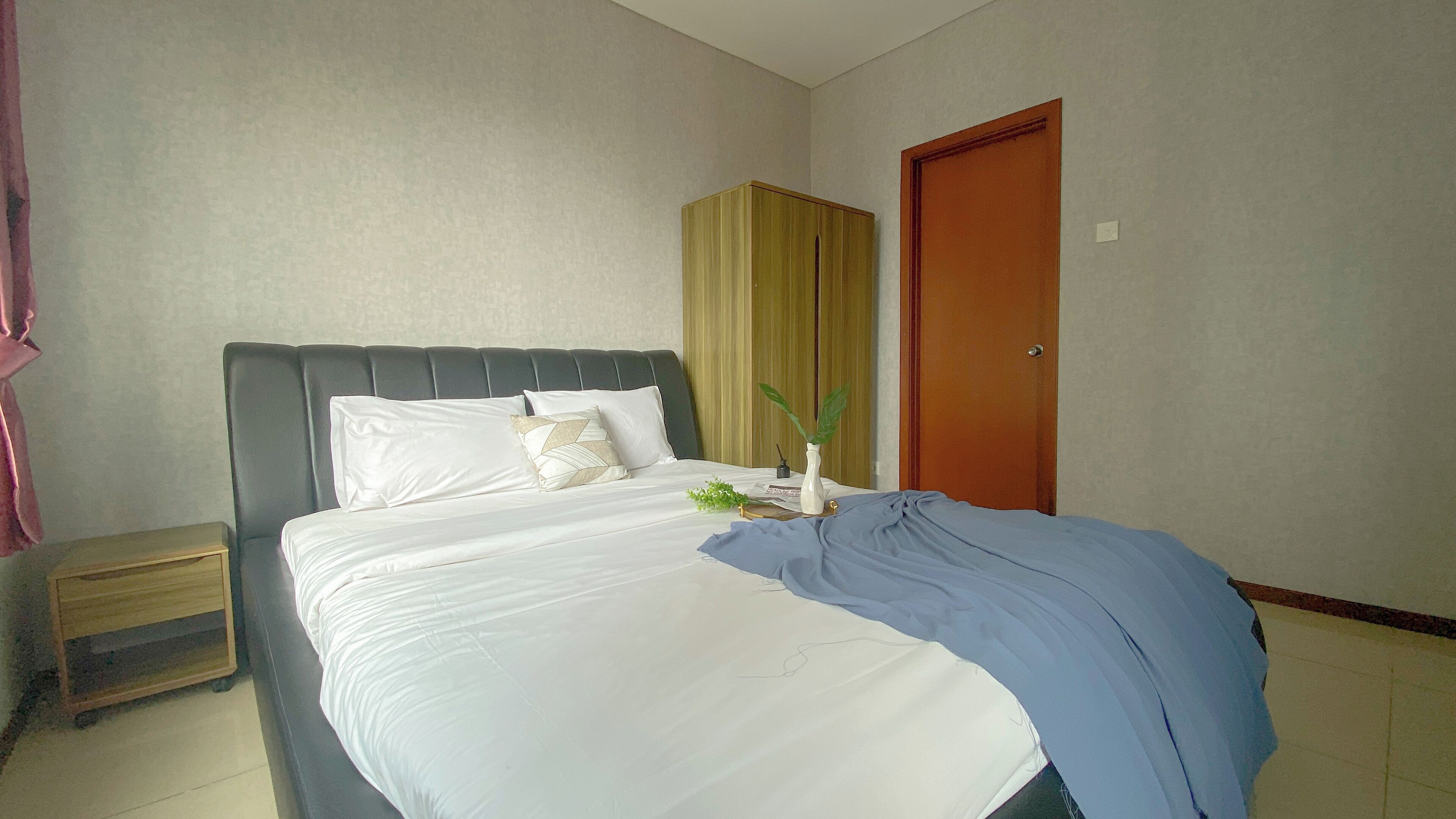 Een net opgemaakt bed met witte lakens en een blauwe deken, een houten nachtkastje met lade, een houten garderobe en een houten deur.