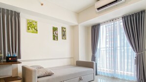 A modern hotel room with a grey sofa, a small table with a bottle and a travel bag, and a window with grey curtains. - Restful And Chic 2Br At Meikarta Apartment (Cikarang)