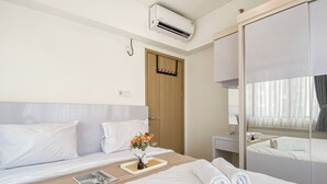 A hotel room with a bed, a bedside table with a vase of flowers, a mirror, and an air conditioning unit. - Restful And Chic 2Br At Meikarta Apartment (Cikarang)