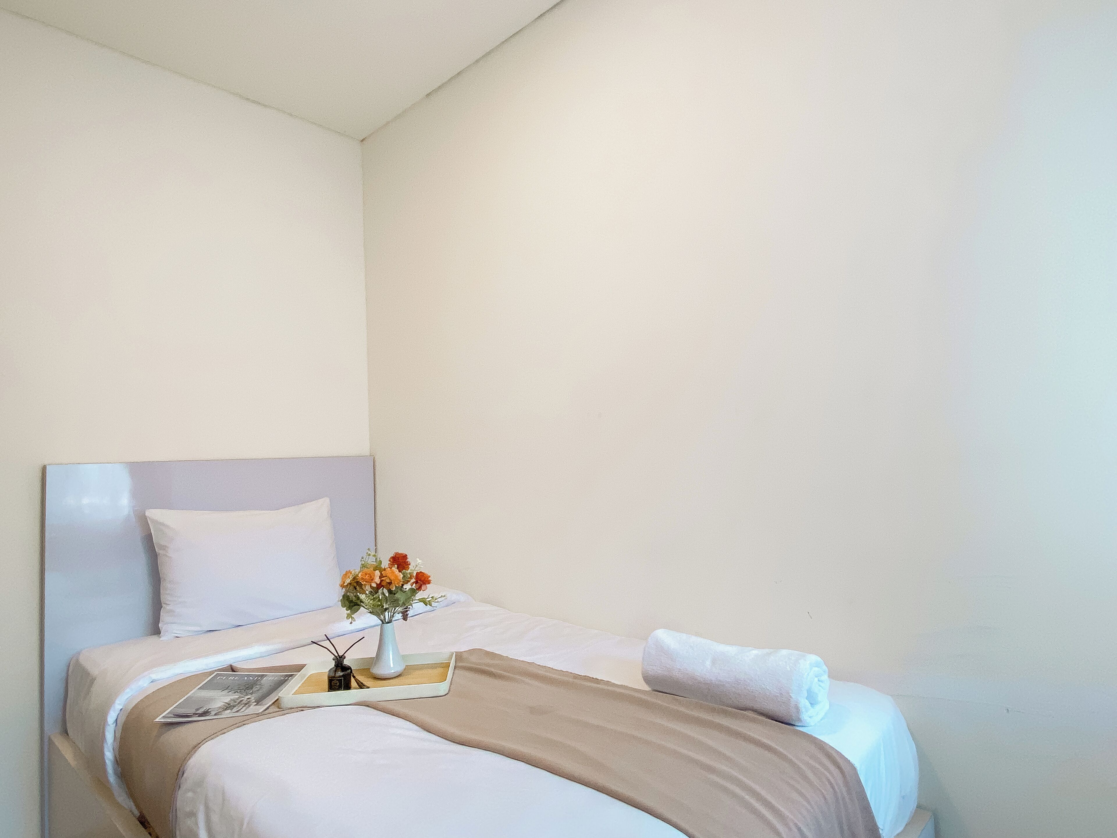Un lit bien fait, avec un oreiller blanc, une couverture beige et une serviette blanche. On peut voir un petit vase avec des fleurs sur le lit.