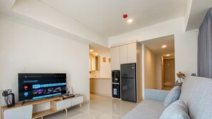 A modern living room with a flat-screen TV, a sofa, a kitchen area with a refrigerator, and a bathroom visible in the background. - Restful And Chic 2Br At Meikarta Apartment (Cikarang)