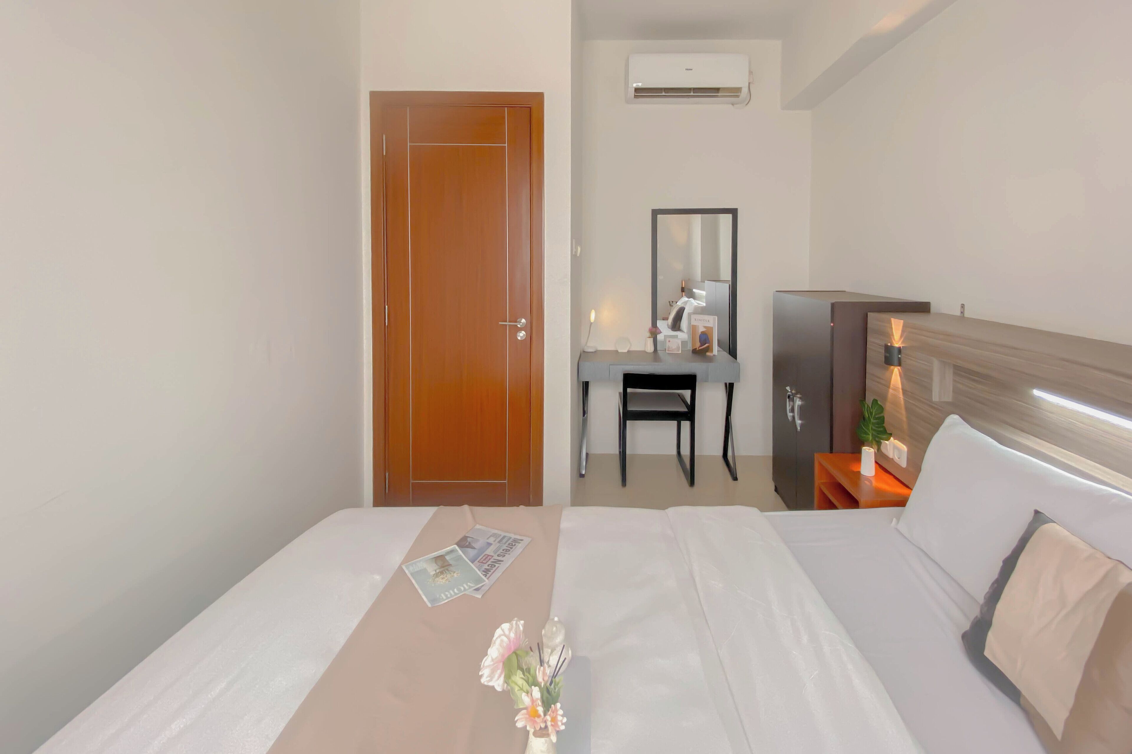A hotel room with a wooden door, a desk with a chair, a mirror, and a bedside table with a lamp.