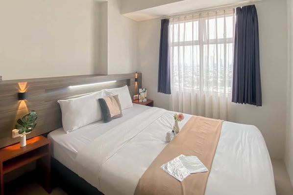 A hotel room with a large bed, a nightstand with a lamp, a window with curtains, and a view of the city.