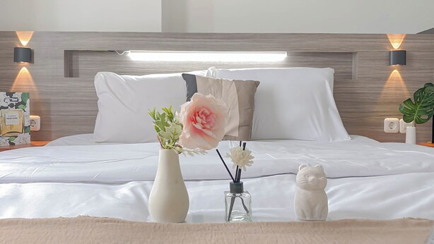 A neatly made bed with white linens, a vase with flowers, a small glass bottle, and a cat figurine on a bedside table. - Warm And Comfortable 1Br Apartment At Vittoria Residence (Jakarta)
