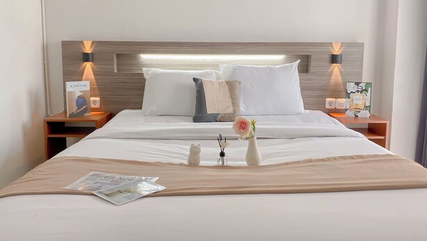 A hotel room with a neatly made bed, a nightstand with a lamp, a vase with flowers, and a magazine on the bed. - Cozy Studio Apartment At Vittoria Residence (Jakarta)