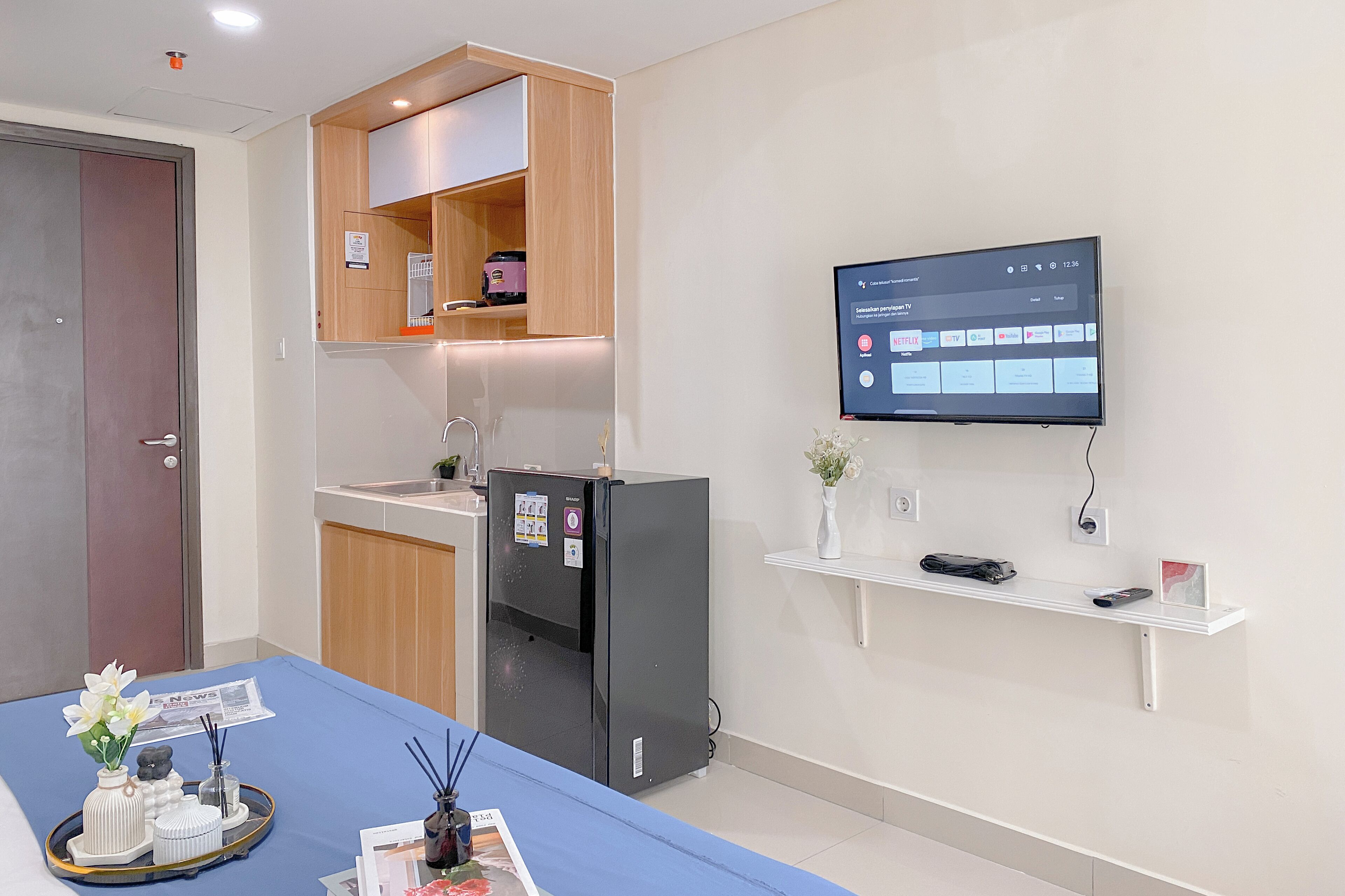 A hotel room with a blue bedspread, a refrigerator, a TV mounted on the wall, and a small shelf with a vase and a picture frame.