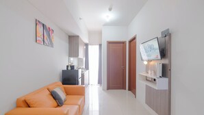 A modern living room with a sofa, a flat-screen TV mounted on a wooden wall, and a kitchen area in the background. - Warm And Comfy 2Br Apartment At Vittoria Residence (Cikarang)