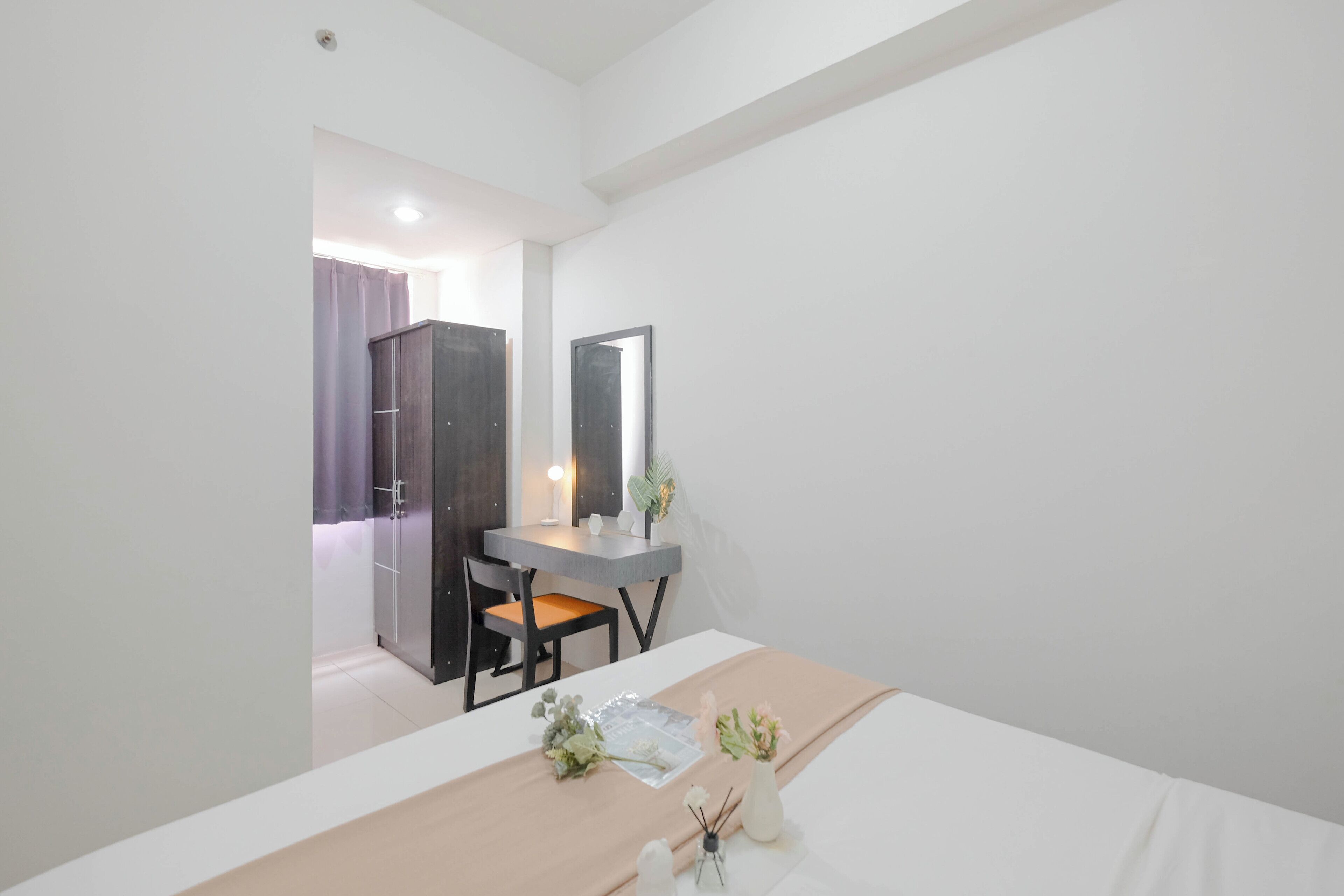 Quarto com cama, escrivaninha e cadeira. Há um espelho na parede e uma porta que dá para outro cômodo.
