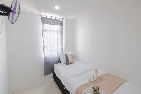 A minimalist bedroom with a bed, a window with curtains, a standing fan, and a small table with a vase. - Warm And Comfy 2Br Apartment At Vittoria Residence (Cikarang)