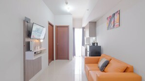 A modern living room with a sofa, a flat-screen TV mounted on a wooden panel, and a kitchen area visible through an open door.