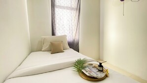 Un petit lit bien fait, avec un matelas blanc et deux oreillers, une fenêtre ornée de rideaux légers et une petite plante posée sur un plateau à côté du lit.