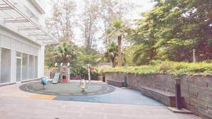Um parquinho com escorrega e baloiços, rodeado por um edifício com grandes janelas e um espaço pavimentado.