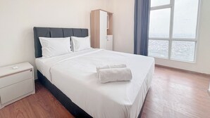 Cama bem arrumada com lençóis brancos e cabeceira cinzenta, uma pequena mesa de cabeceira e vista para a cidade através de uma janela grande.