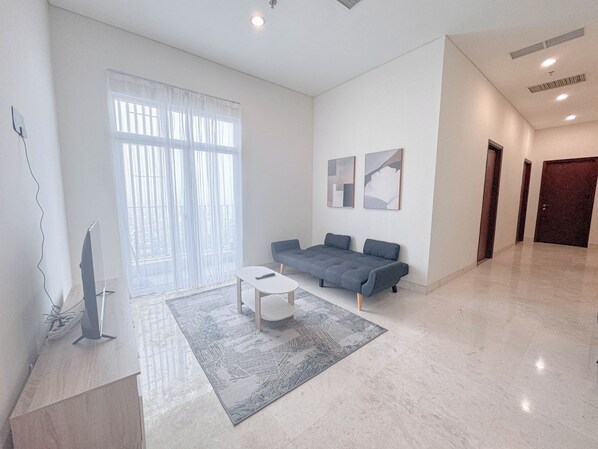 A spacious living room with a grey sofa, a white coffee table, and a large window with sheer curtains. - Homey And Simply 3Br At Grand Sungkono Lagoon Apartment (Surabaya)