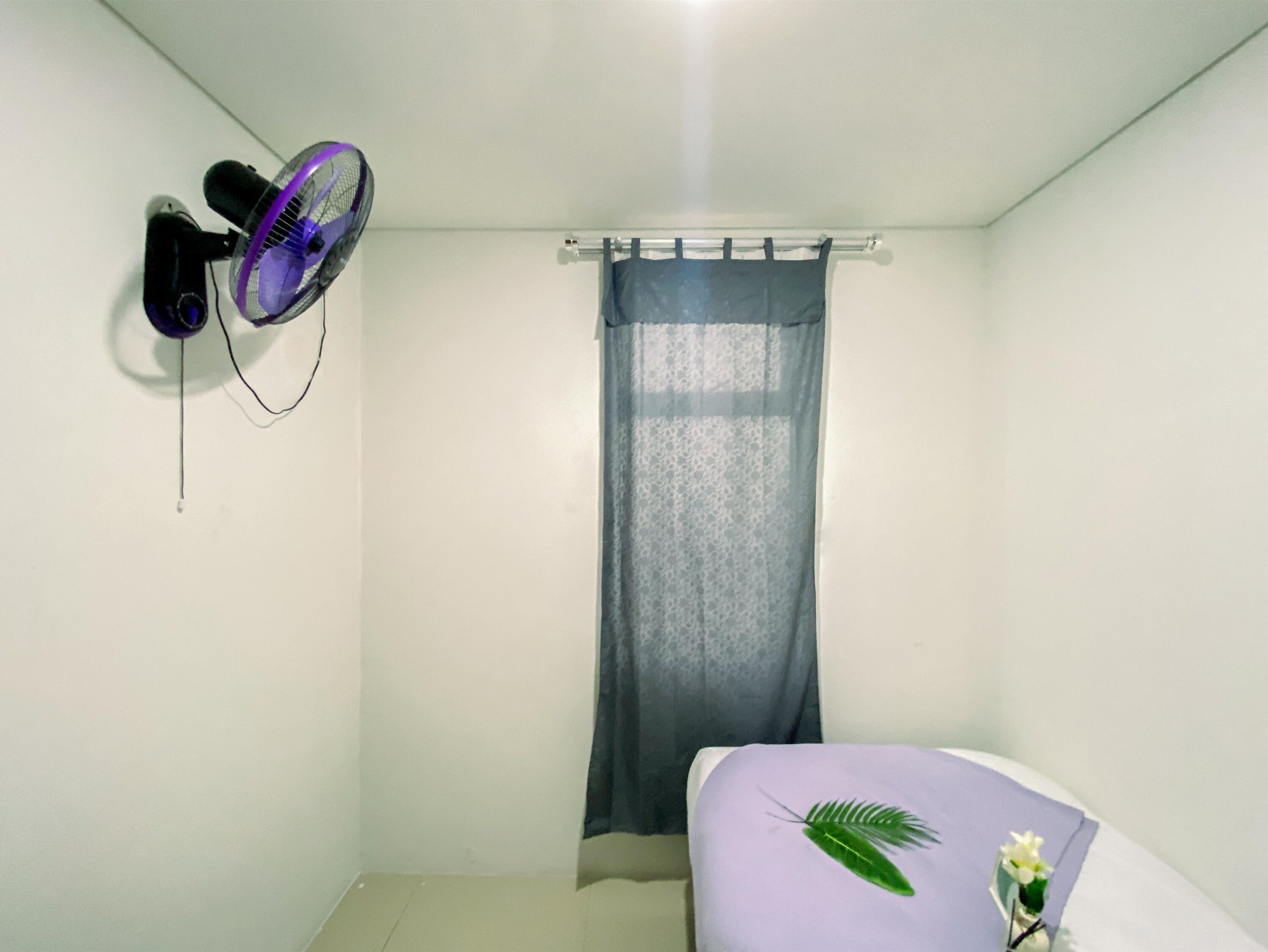 A small, clean room with a bed, a wall-mounted fan, and a curtain.