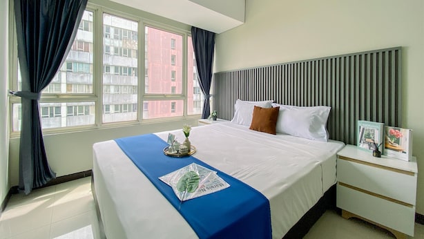 A hotel room with a large bed, a nightstand, a window with curtains, and a view of buildings outside. - Full Furnished And Homey 3Br Apartment Nifarro Park (Jakarta)