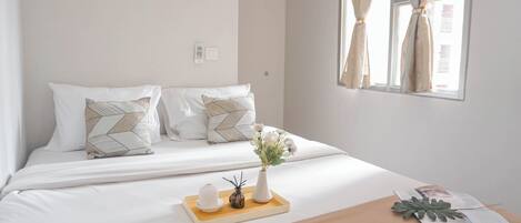 A neatly made bed with white bedding and patterned pillows, a tray with a vase of flowers, and a window with curtains.