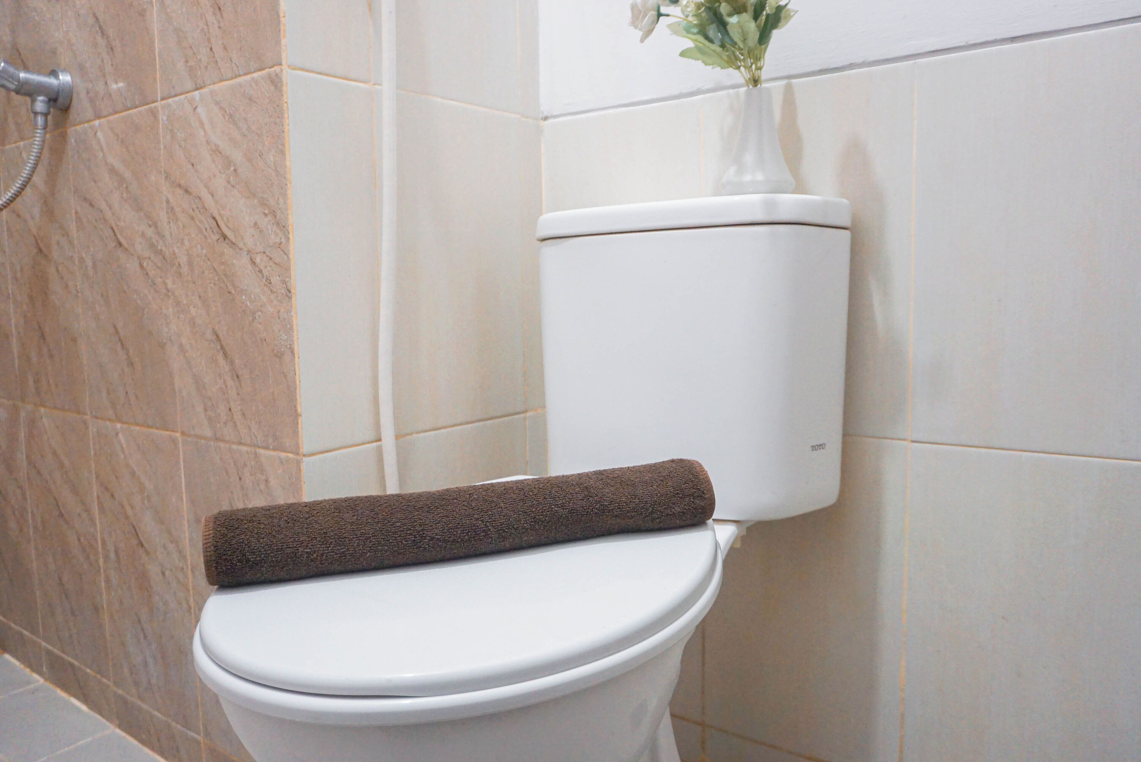 Eine Toilette mit einem braunen Handtuch auf dem Sitz, einer Duschbrause links und einer Vase mit Blumen an der Wand.