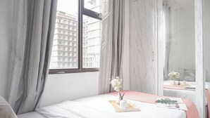 Ett hotellrum med en säng, ett fönster med gardiner, en vas med blommor och en spegel.