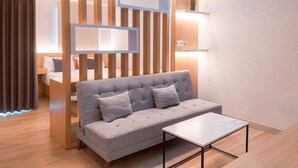 A modern living room with a grey sofa, a small table, wooden slats, and a wall-mounted shelf with a plant. - Cozy Studio Apartment Parkland Avenue (South Tangerang)
