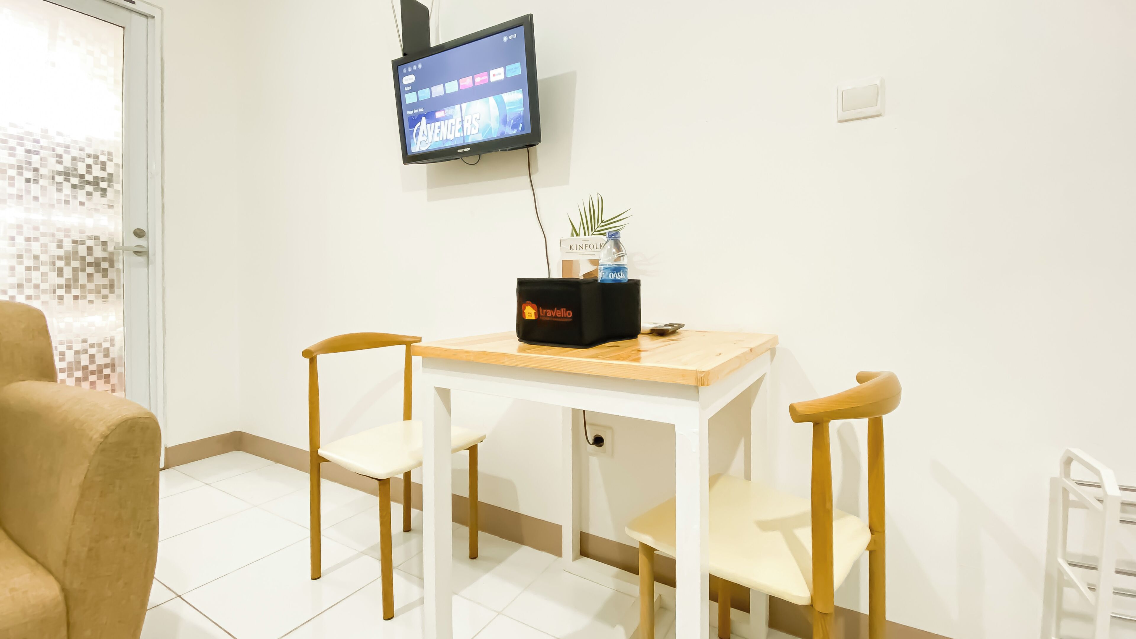 A hotel room with a white tiled floor, a wooden table with two chairs, a television mounted on the wall, and a small plant on the table.
