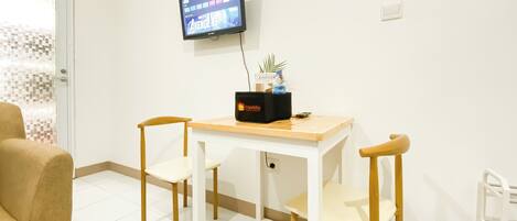 A hotel room with a white tiled floor, a wooden table with two chairs, a television mounted on the wall, and a small plant on the table.