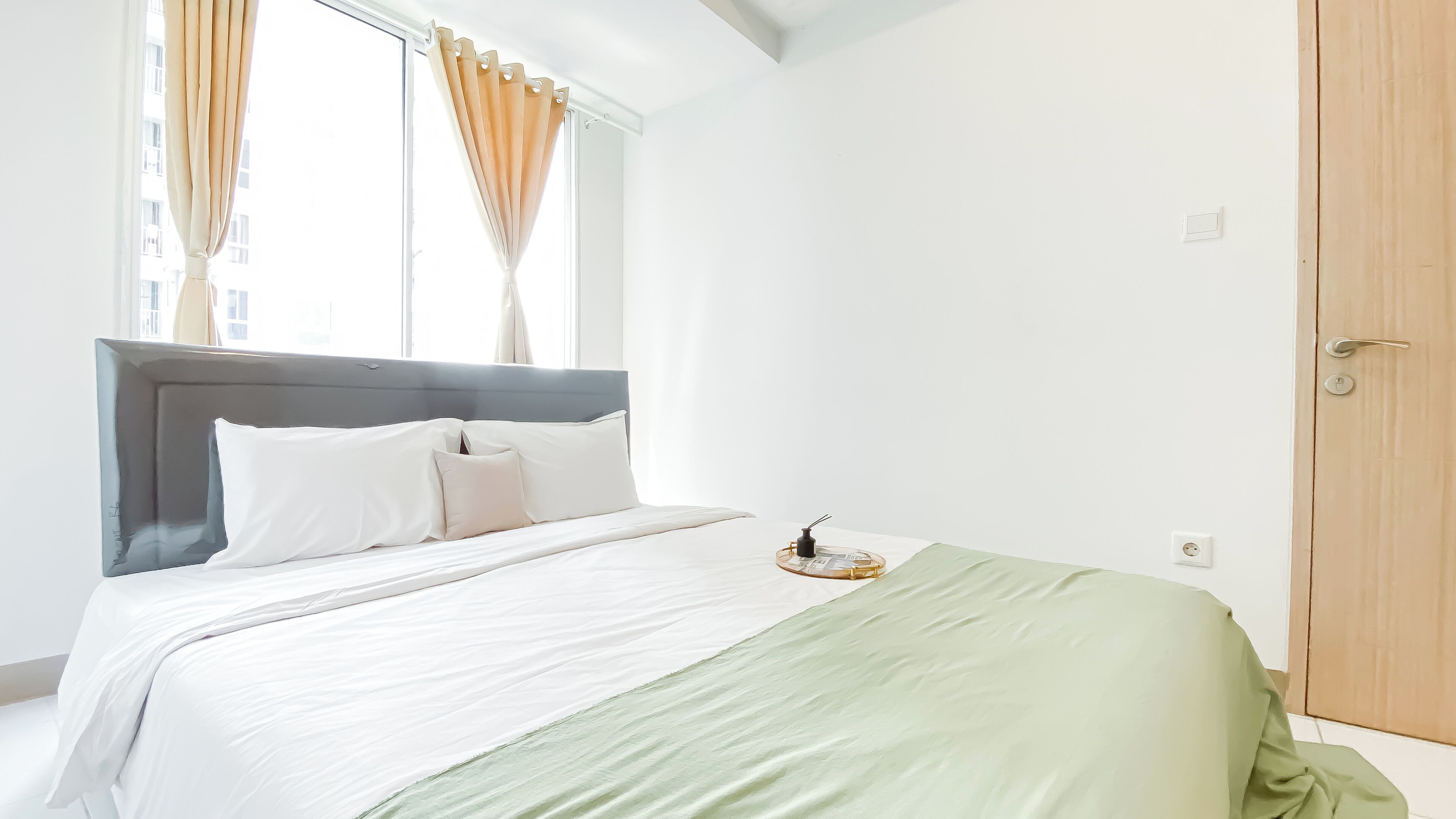 A neatly made bed with white linens and a green throw blanket, a bedside table with a lamp, and a window with curtains.