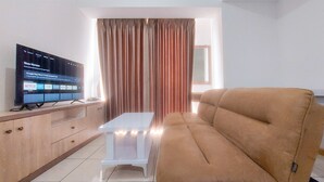 A living room with a beige sofa, a flat-screen TV on a wooden cabinet, a small white table, and a window with curtains. - Comfy 1Br Apartment At M-Town Residence (Gading Serpong)