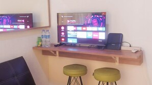 A room with a TV on a shelf, two green stools, and a black couch. - Lovely 2Br Apartment At Belmont Residence Puri (Jakarta)