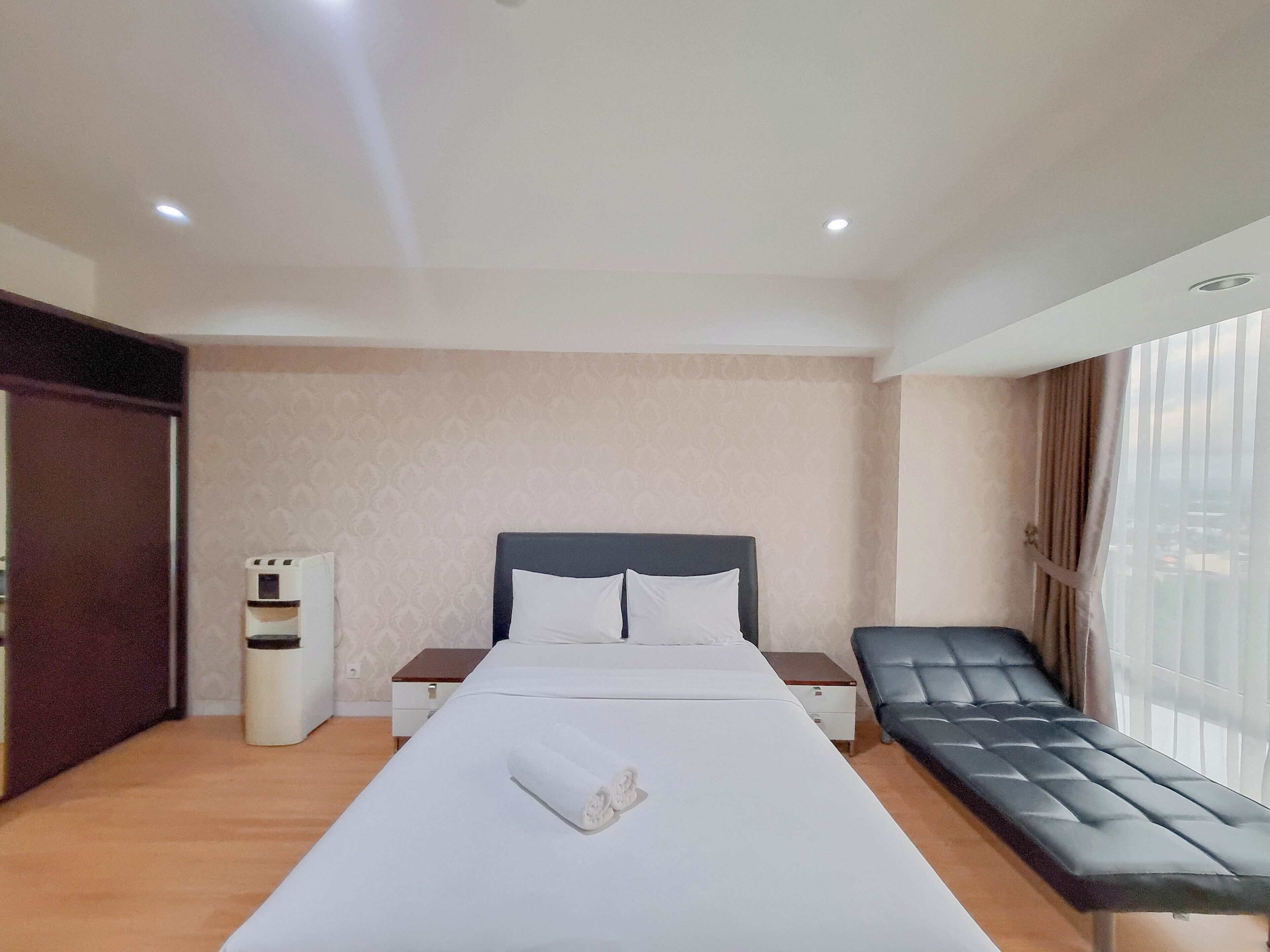 A hotel room with a large bed, a bedside table, a small bedside table, a bench, a water dispenser, and a window with curtains.