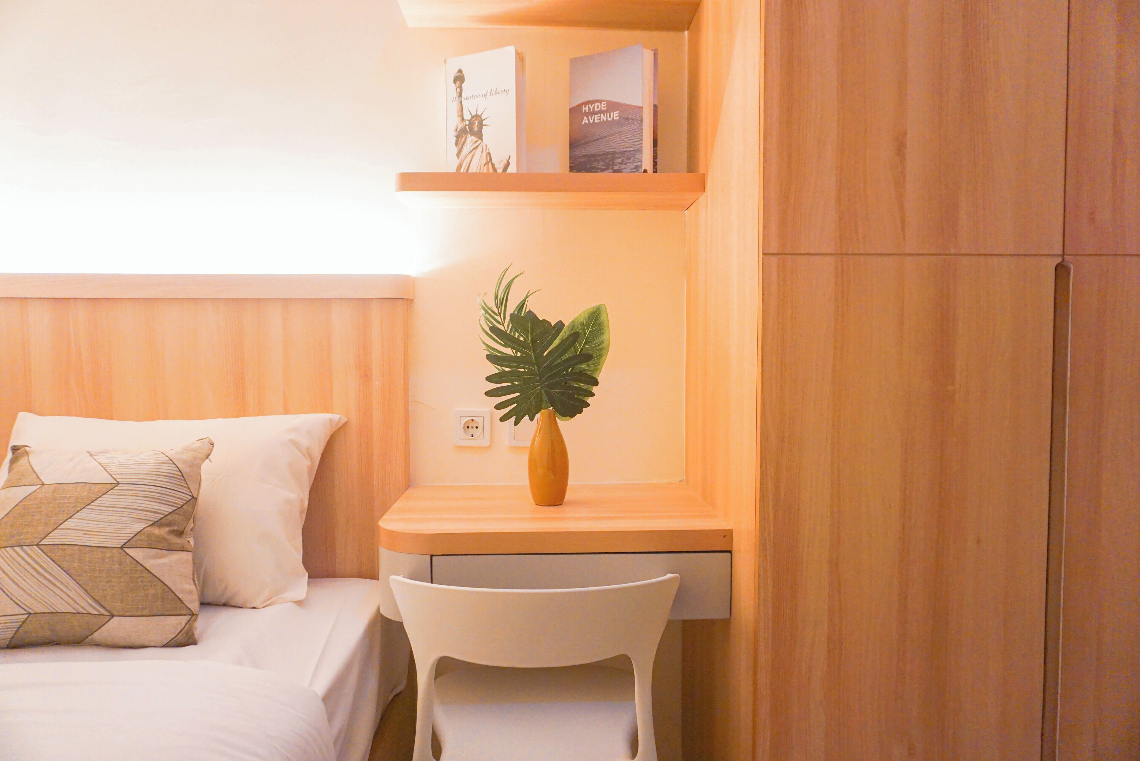 Un dormitorio ordenado con cama, una silla, un escritorio pequeño y una estantería con libros y una planta.