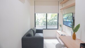 Un espacio de oficina moderno con un sofá gris, un escritorio de madera, un televisor de pantalla plana y un ventanal con vistas a zonas verdes.