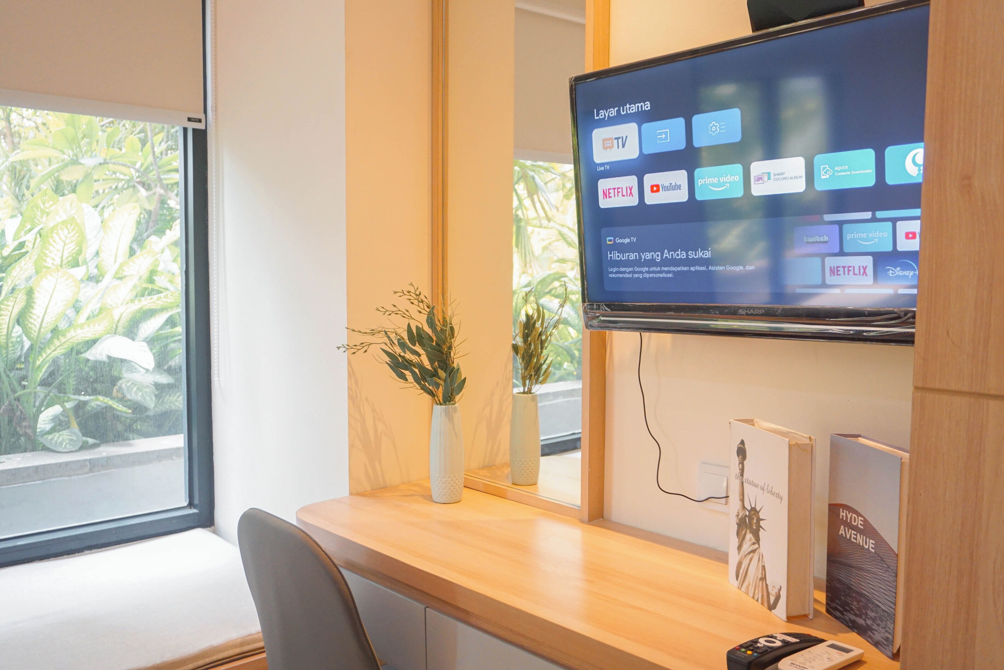 Een moderne hotelkamer met een grote flatscreen televisie aan de muur, waarop diverse streamingdiensten worden getoond. Er is een houten bureau met een bloemenvaas en een lijst met een afbeelding van de Vrijheidsbeeld.