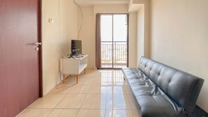 A hallway with a black leather sofa, a white desk with a TV, and a sliding glass door leading to a balcony. - Scenic View 2Br At Apartment Tamansari Panoramic (Bandung)