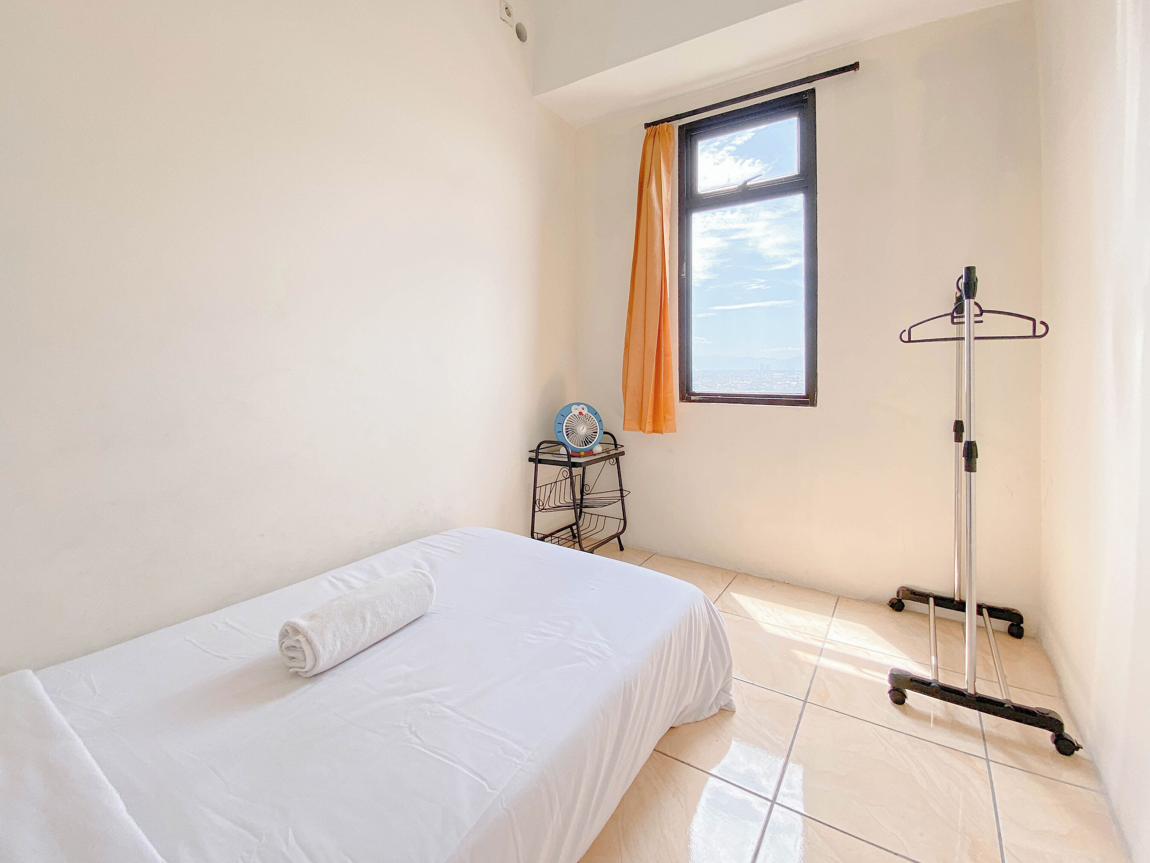 A small, clean room with a single bed, a window with an orange curtain, a wall-mounted fan, and a coat rack.