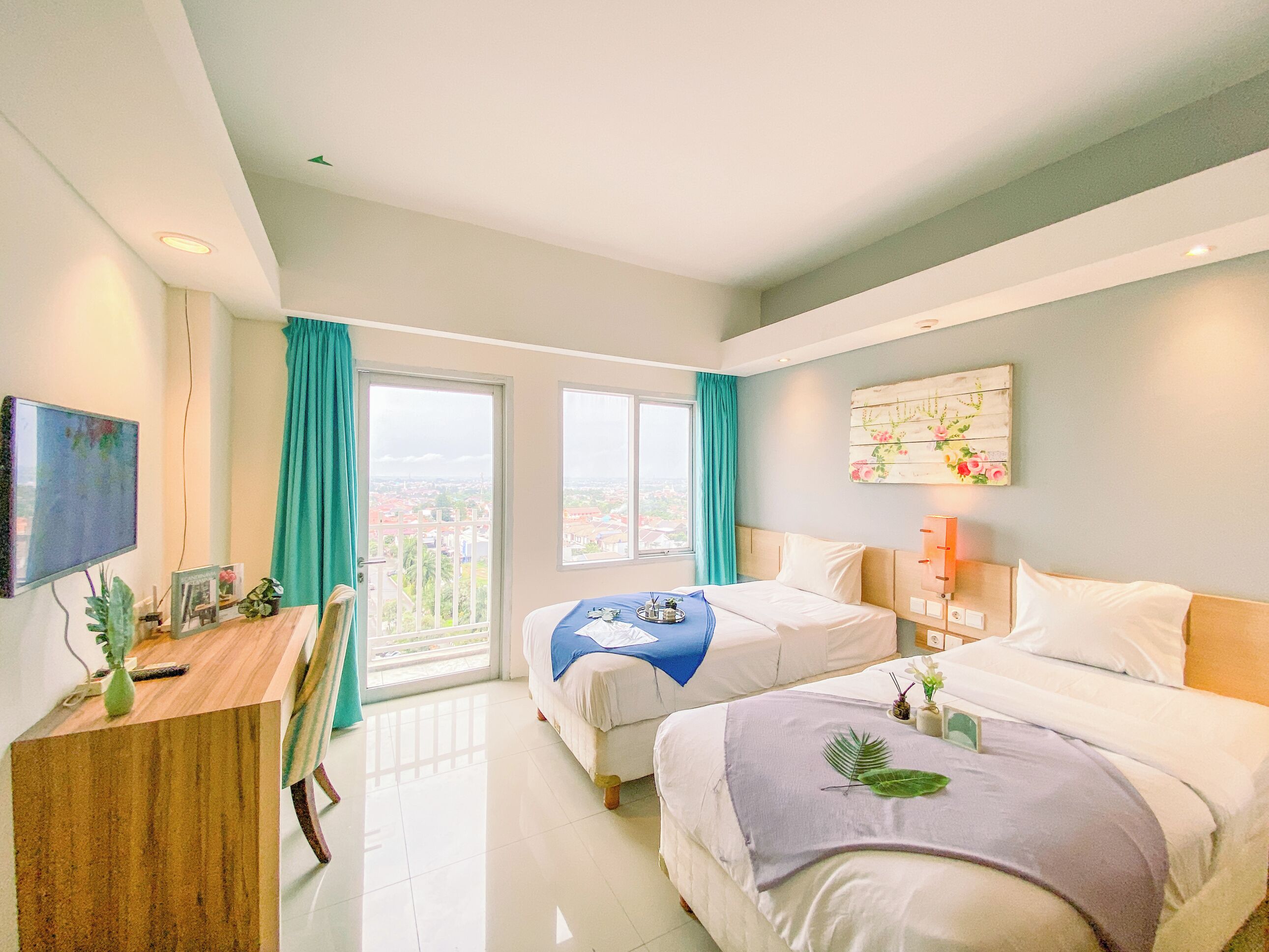 A hotel room with two beds, a desk, a TV, and a balcony with a city view.