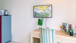 Habitació amb televisió de pantalla plana penjada a la paret, escriptori de fusta, cadira, nevera petita i una planta en test.