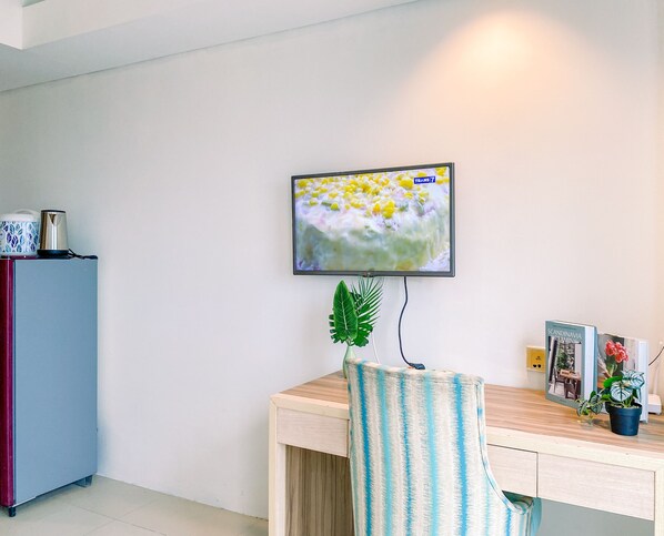 A room with a flat-screen TV mounted on the wall, a wooden desk, a chair, a small refrigerator, and a potted plant. - Comfortable Studio (No Kitchen) At Bogor Icon Apartment (Bogor)
